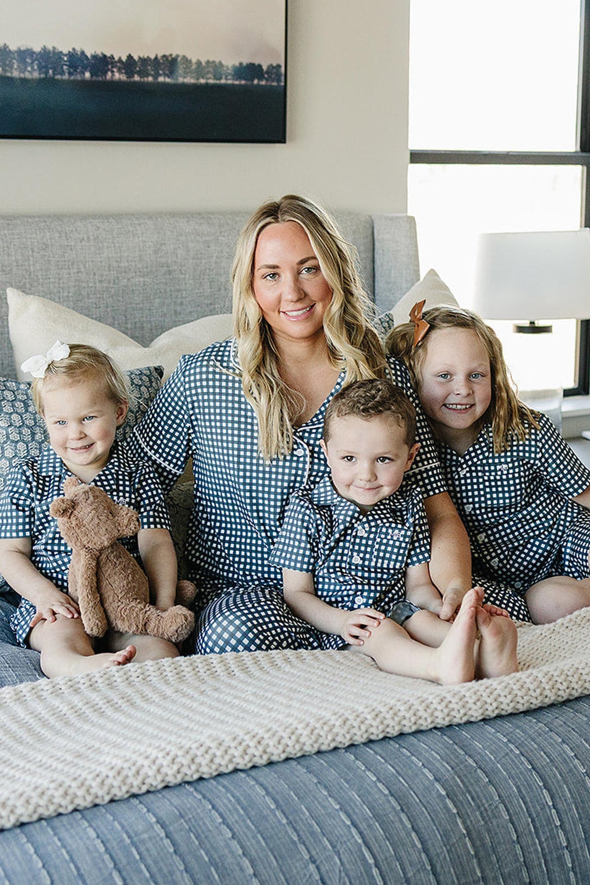 A woman and three children sit on a bed wearing matching navy gingham pajamas, smiling warmly at the camera.