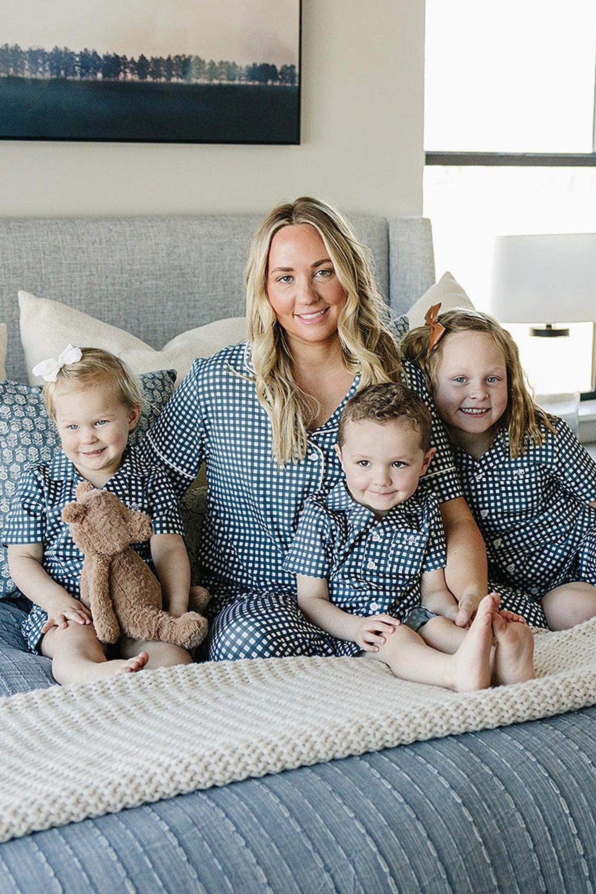 A woman and three children sit on a bed wearing matching navy gingham pajamas, one child holding a teddy bear.