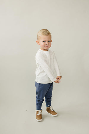 A young child stands confidently in a light grey long-sleeve shirt and navy joggers, paired with brown high-top shoes.