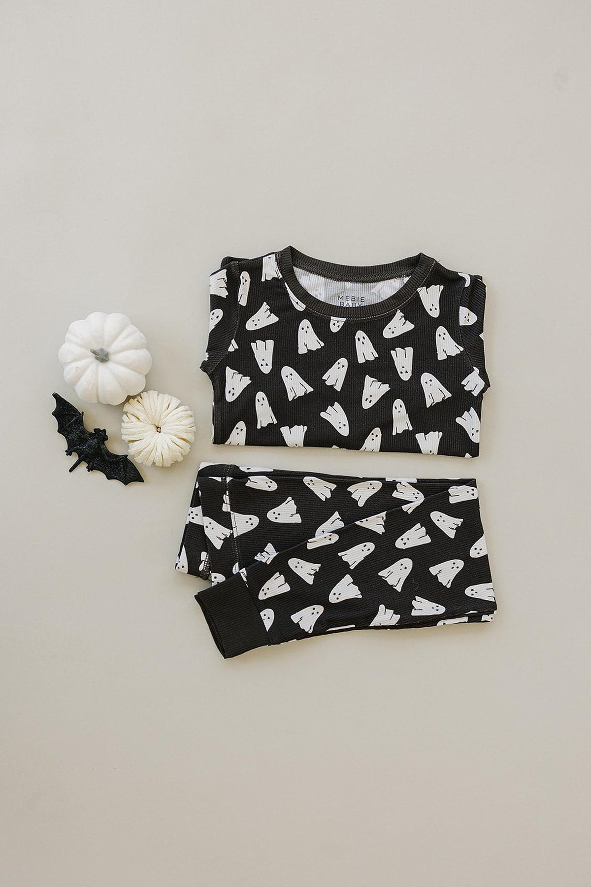Black pajama set with white ghost print, displayed with two white pumpkins and a small black bat decoration.