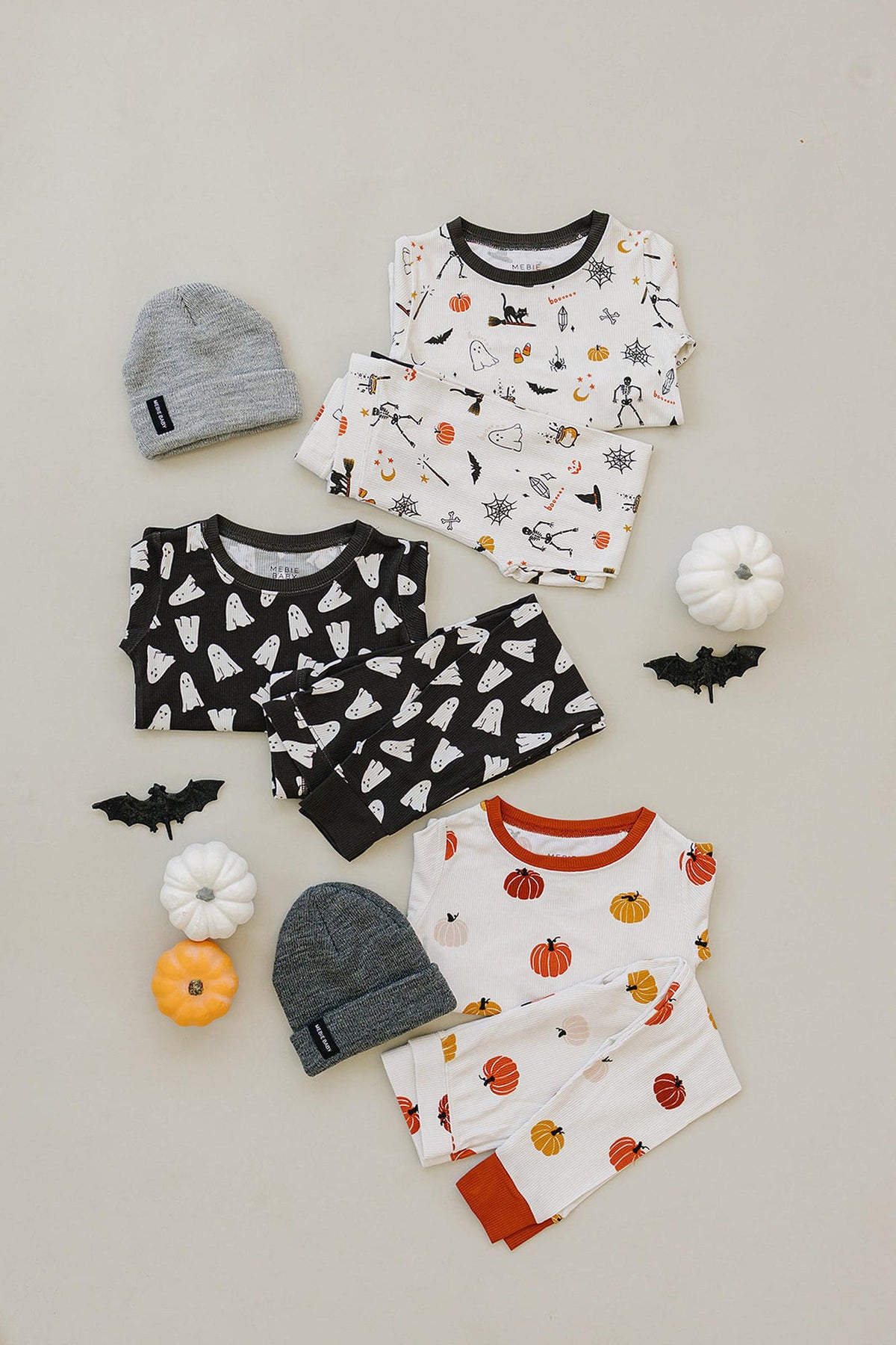 Three sets of Halloween-themed pajamas with ghost and pumpkin prints, accompanied by gray hats and decorative pumpkins.