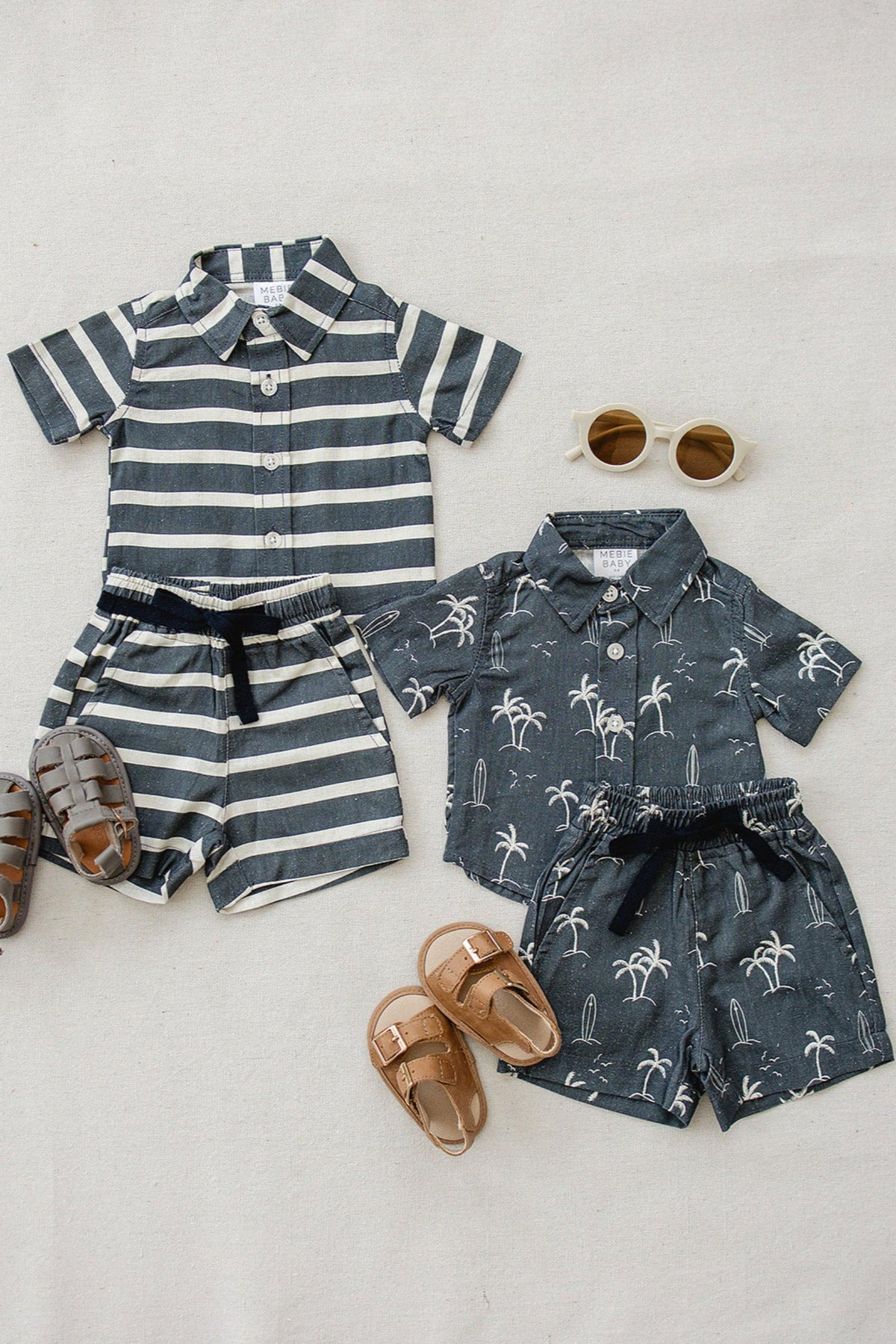 Two sets of childrens clothing include navy striped and palm tree print shirts with matching shorts, plus sandals.