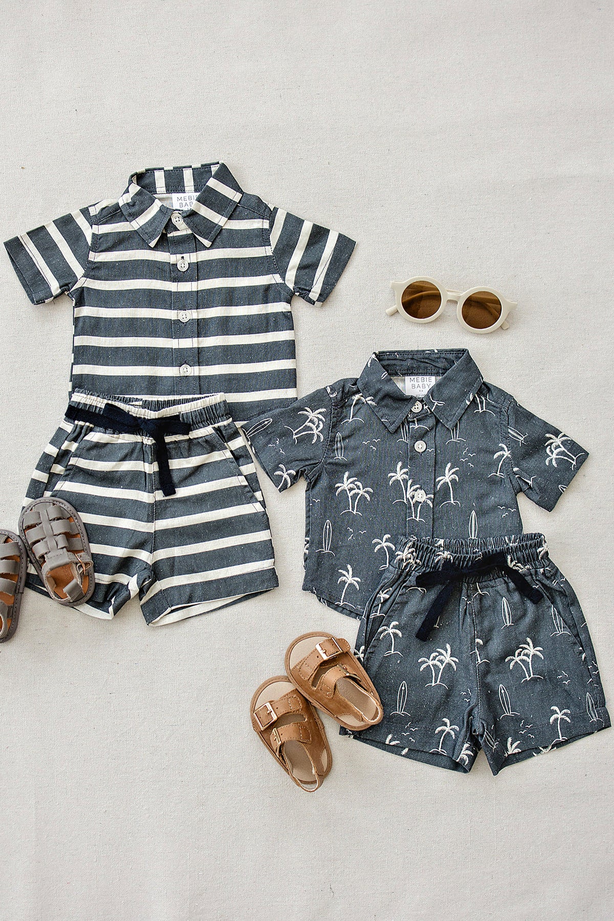 Two Mebie Baby outfits with navy stripes and palm prints, paired with sandals and sunglasses on a beige background.