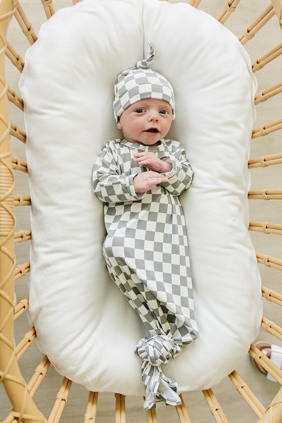 A baby wearing a light green checkered knot gown and matching hat lies on a white cushioned surface.