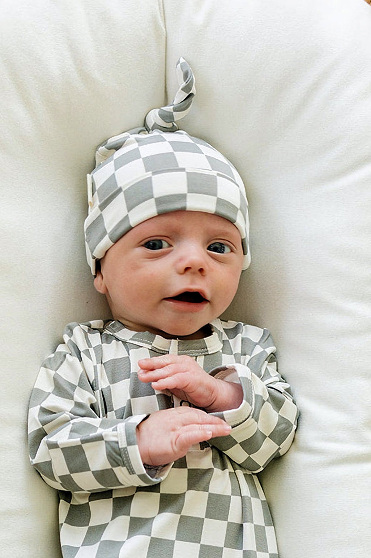 A newborn wearing a light green checkered knot hat and matching outfit lies on a soft white surface.