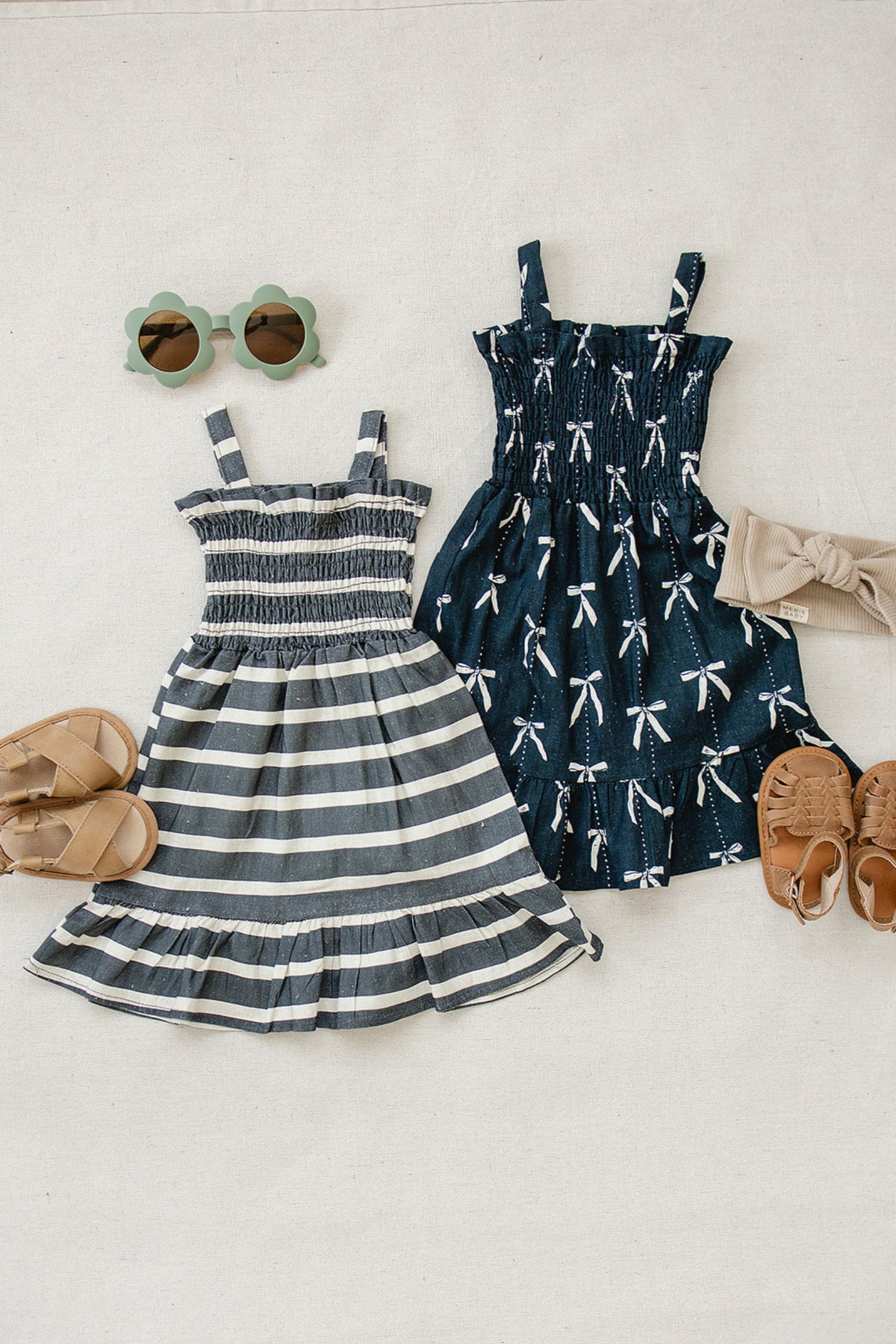 Two childrens dresses, one navy with bows and one striped, are displayed with sandals, sunglasses, and a headband.