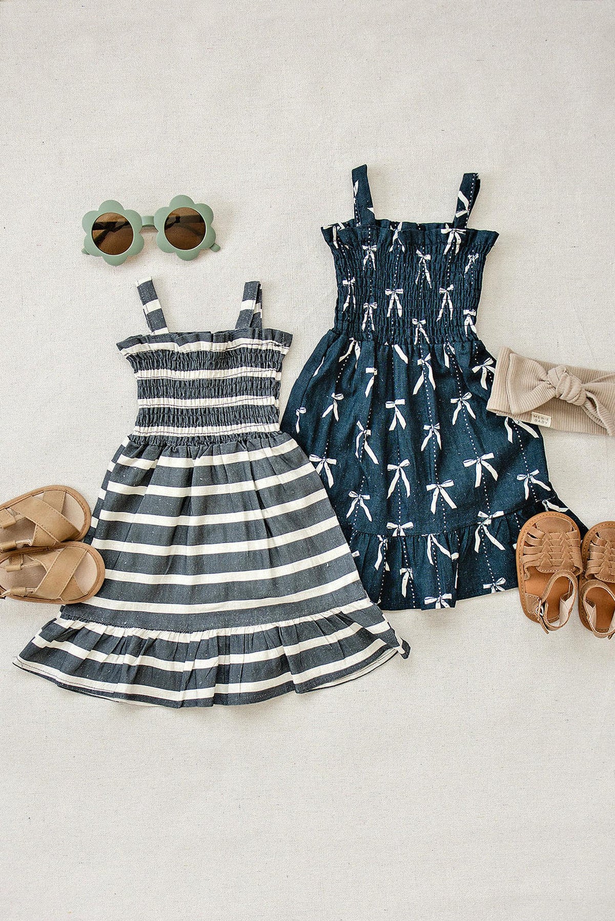 Two summer dresses, one navy with bow print and another striped, paired with sandals, sunglasses, and a headband.
