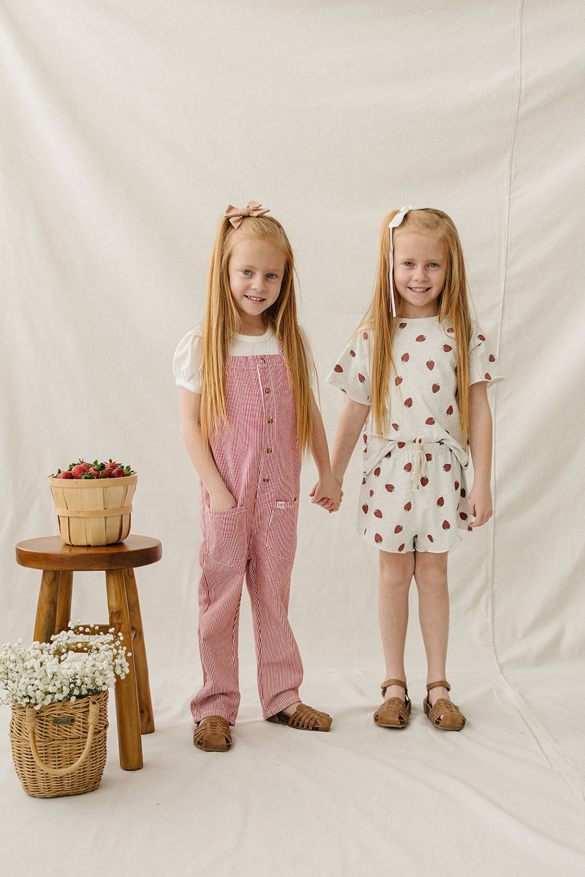 Two young girls stand side by side, one in a pink striped jumpsuit and the other in a light grey.