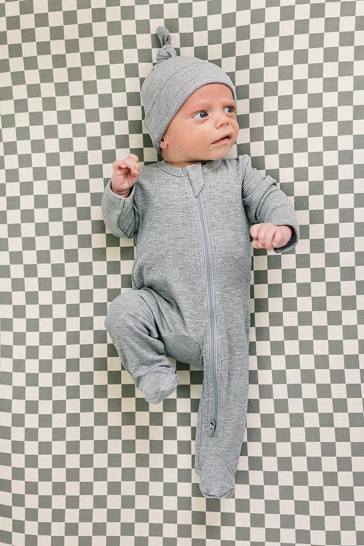 Baby in a heather grey ribbed zip-up pajama with matching hat, lying on a checkered surface.