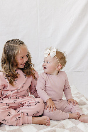 Two children sit on a soft, checkered blanket, wearing matching dusty pink outfits. The older child dons a hoodie and pants with a playful design, while the baby wears a.