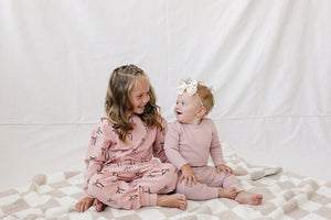 Two young children sit on a soft, checkered blanket, wearing matching blush-colored outfits adorned with bow prints.