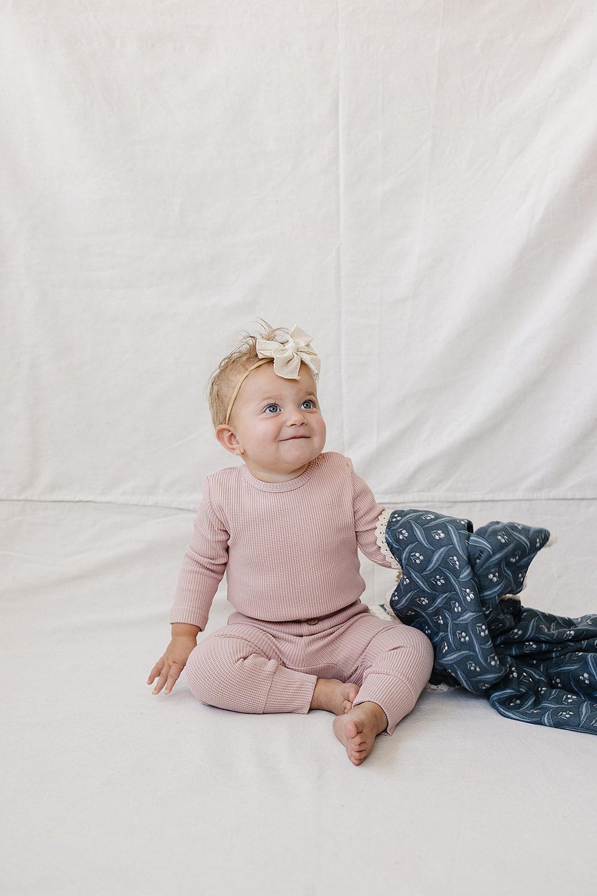 A joyful baby sits on a light fabric backdrop, wearing a dusty pink waffle knit cotton outfit. She has a large bow in her hair and holds a patterned blanket.