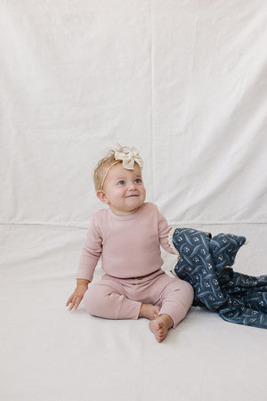 A joyful baby sits on a light fabric backdrop, wearing a dusty pink waffle knit cotton outfit. She has a large bow in her hair and holds a patterned blanket.