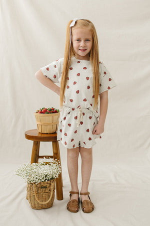 A young girl wears a light grey cotton shorts set with a strawberry print, standing next to a wooden stool.