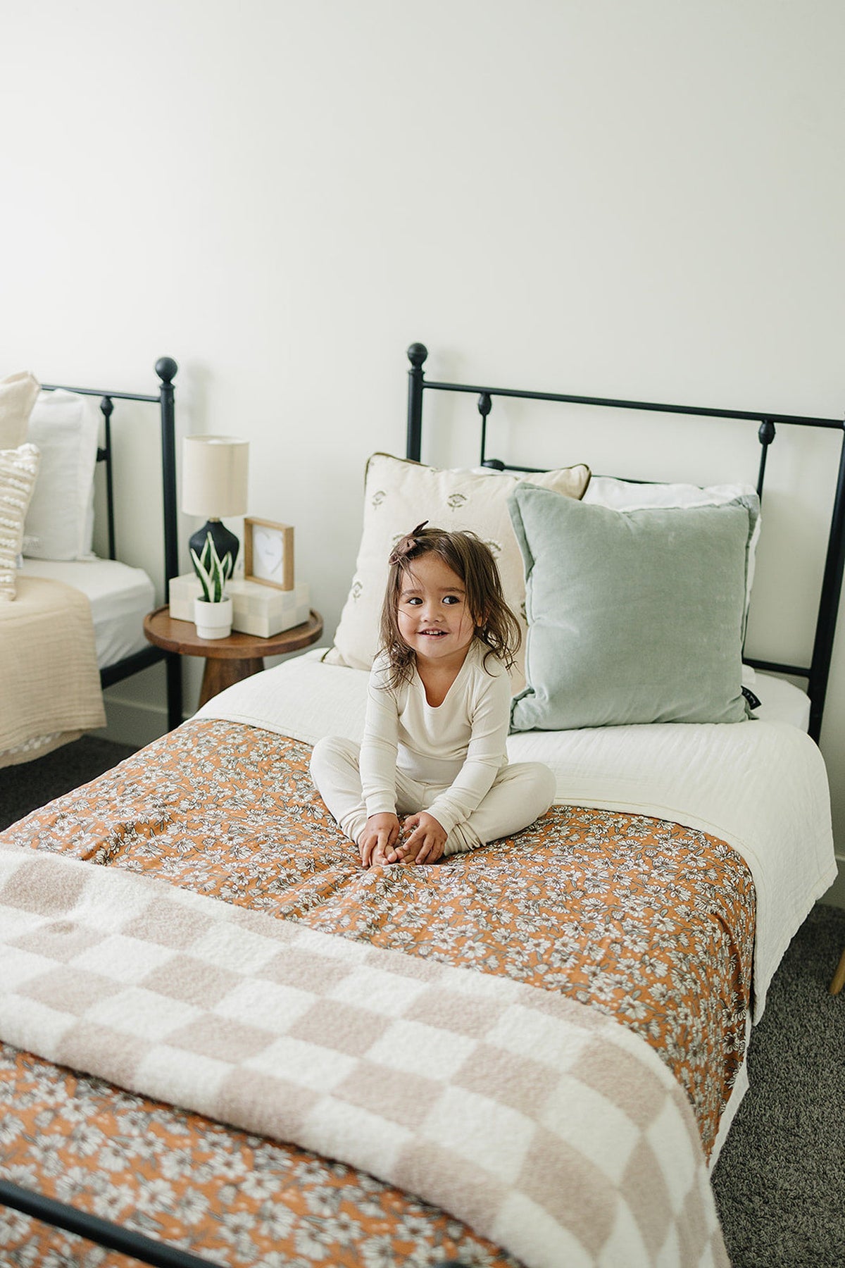 A smiling child sits cross-legged on a bed with a floral quilt, surrounded by decorative pillows and soft furnishings.