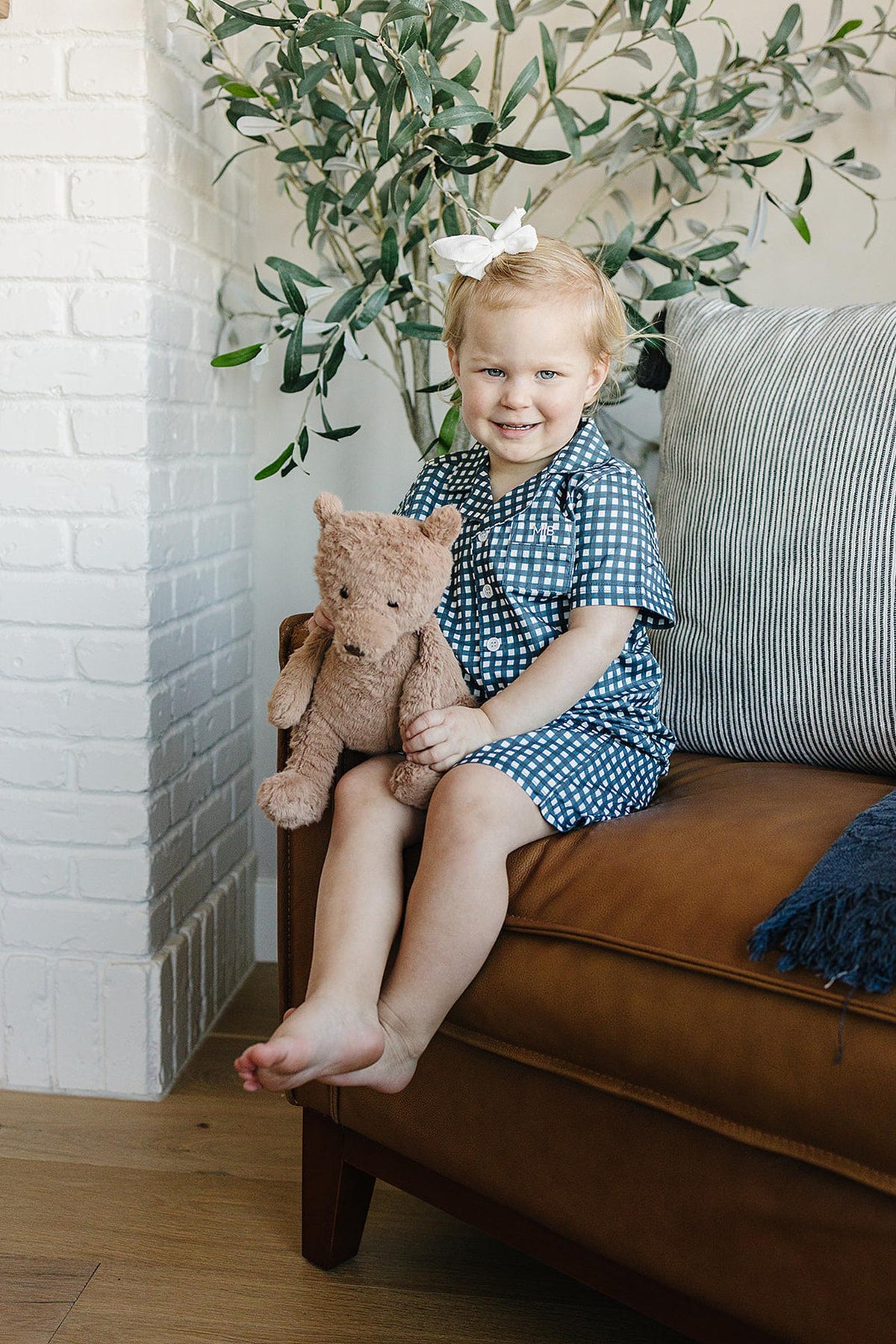 A child in navy gingham pajamas sits on a brown couch, holding a teddy bear, near a plant.
