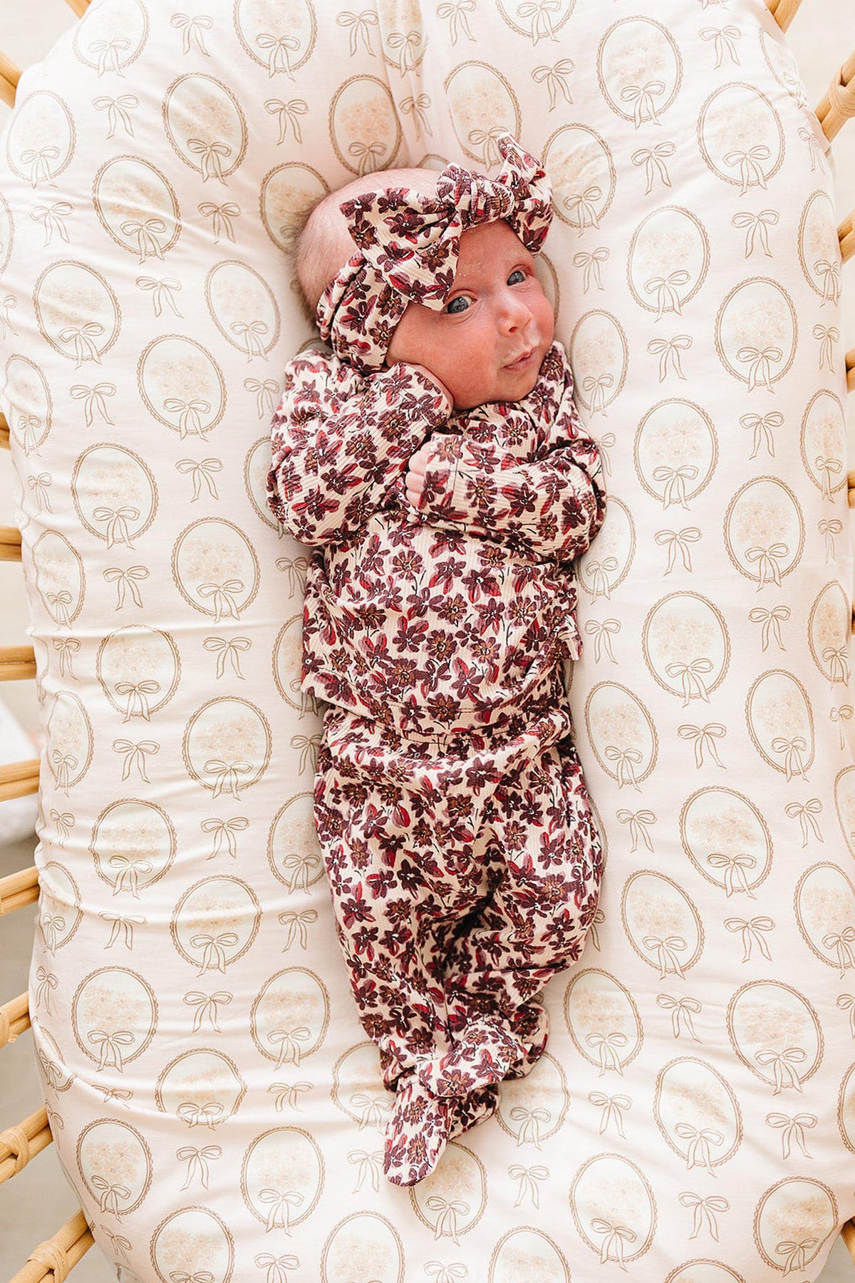 A newborn is comfortably nestled in a bamboo crib, wearing a floral-patterned outfit and matching head wrap. The soft fabric and gentle colors offer a cozy, nurturing environment.