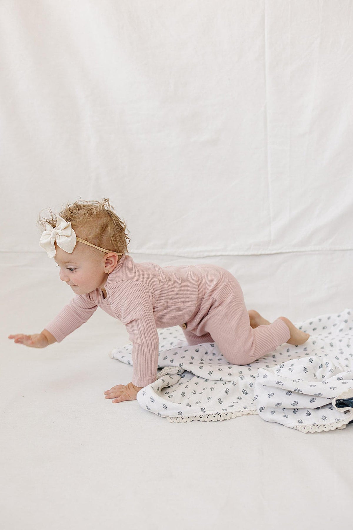 A baby crawls on a soft blanket, wearing a dusty pink waffle knit long sleeve bodysuit and matching pants.