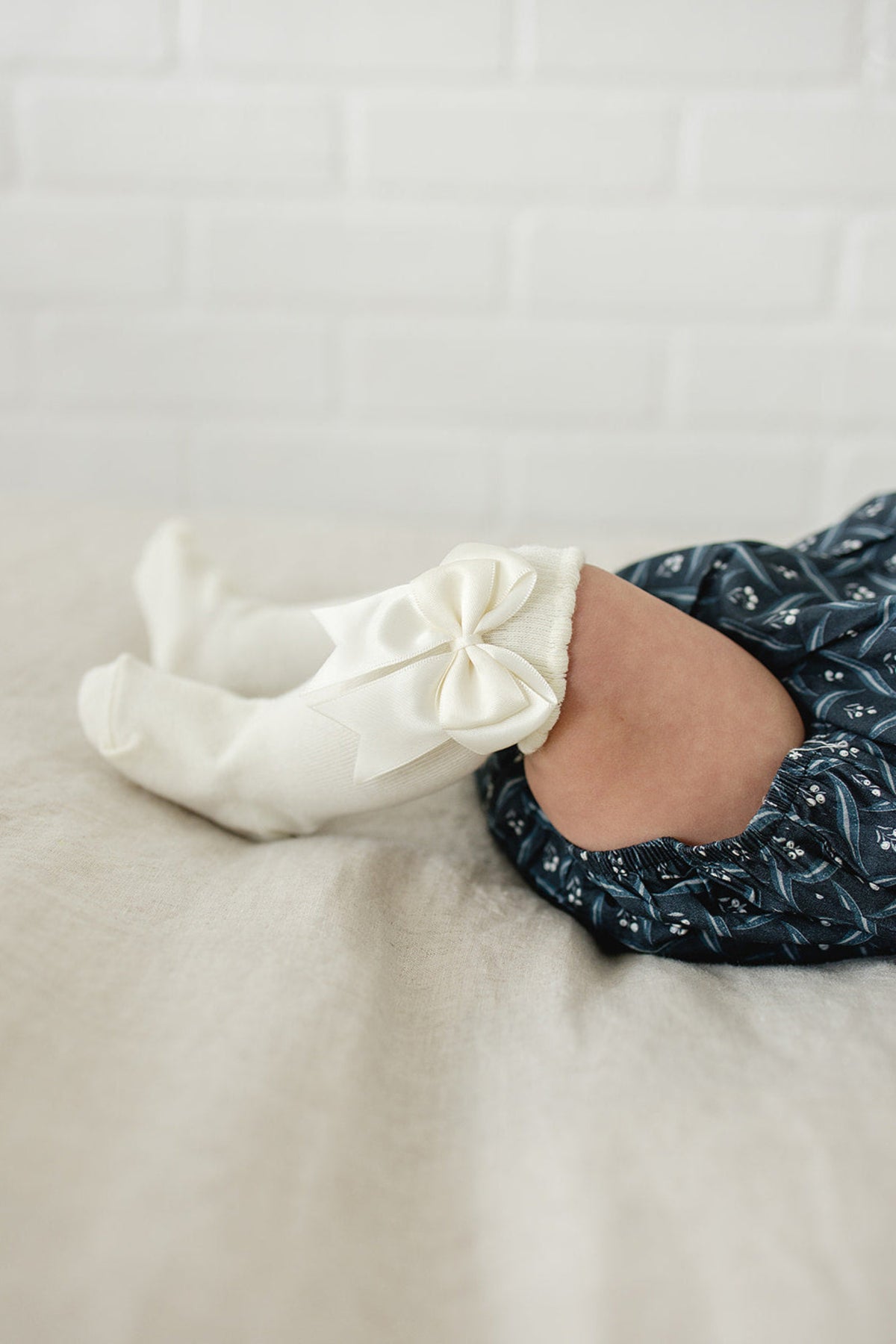 A baby wears cream-colored high-rise socks with bows, paired with a blue patterned outfit on a beige surface.