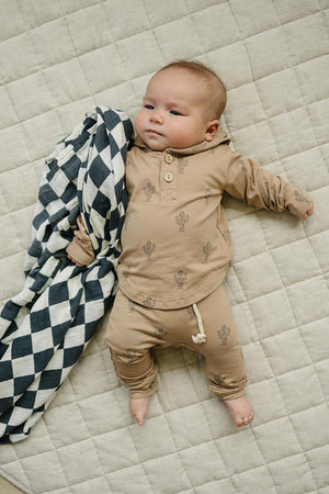 A baby wearing a tan cactus-patterned hooded set lies on a quilted surface with a black and white blanket.
