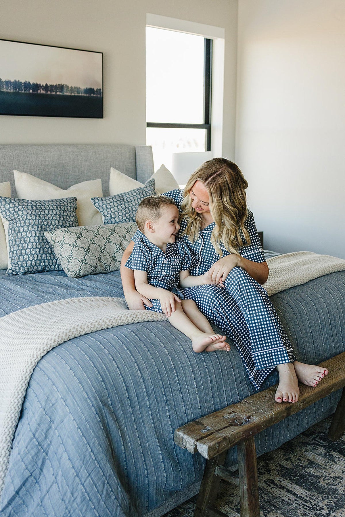 A woman and child wearing matching navy gingham pajamas sit on a bed with blue and white pillows.