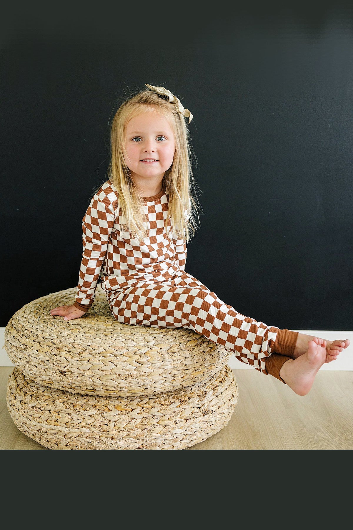 A child wearing a rust checkered pajama set sits on woven cushions against a dark background.