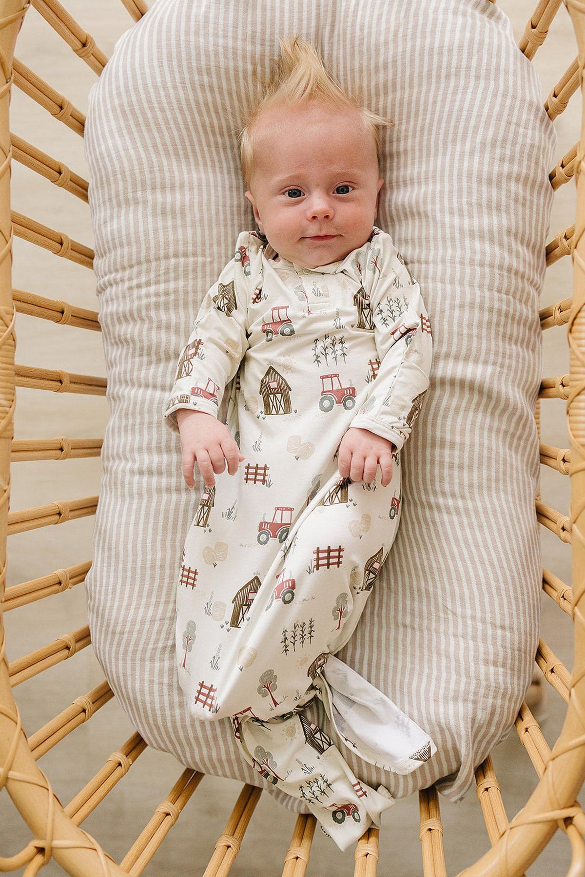 A baby lies comfortably in a woven bassinet, wearing a soft, cream-colored bamboo gown adorned with whimsical farm-themed illustrations. The gown features fold-over hands for added warmth.