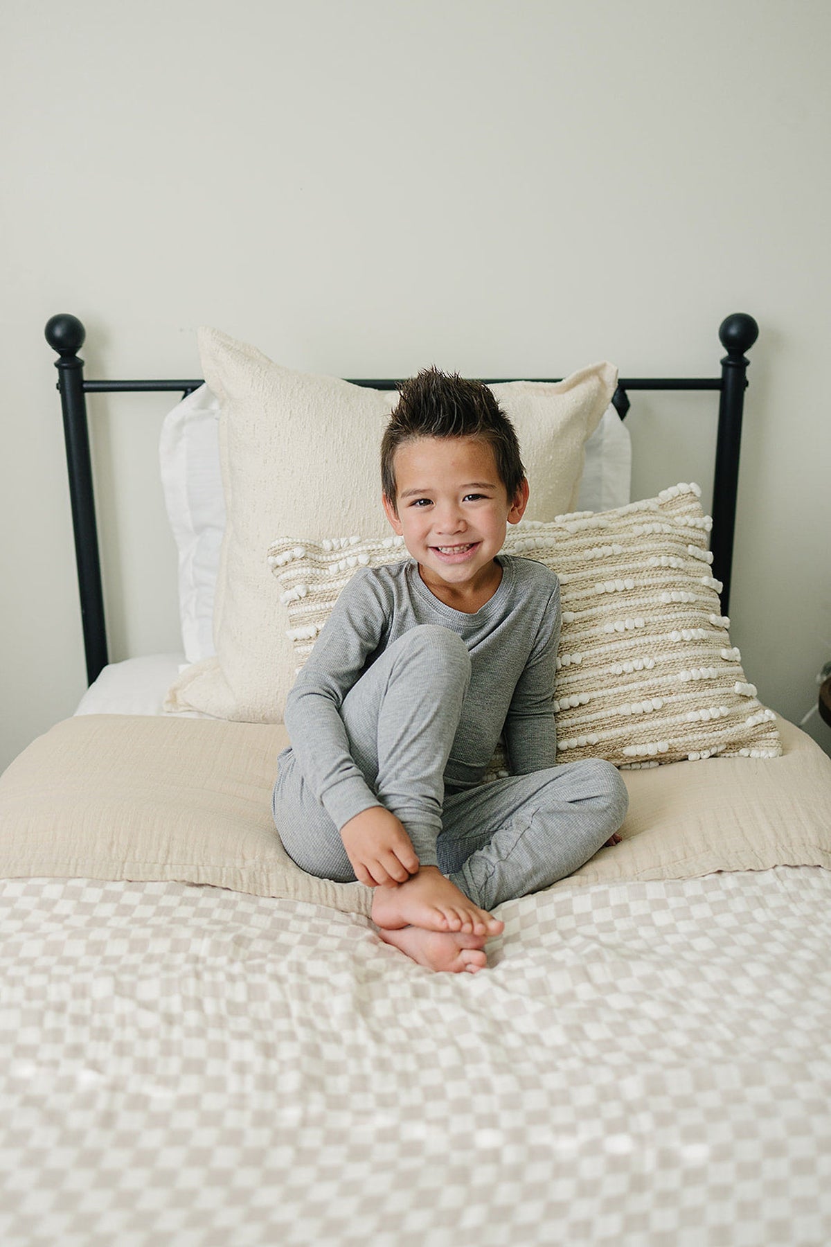 A child in gray pajamas sits on a bed with a taupe checkered quilt and decorative pillows.