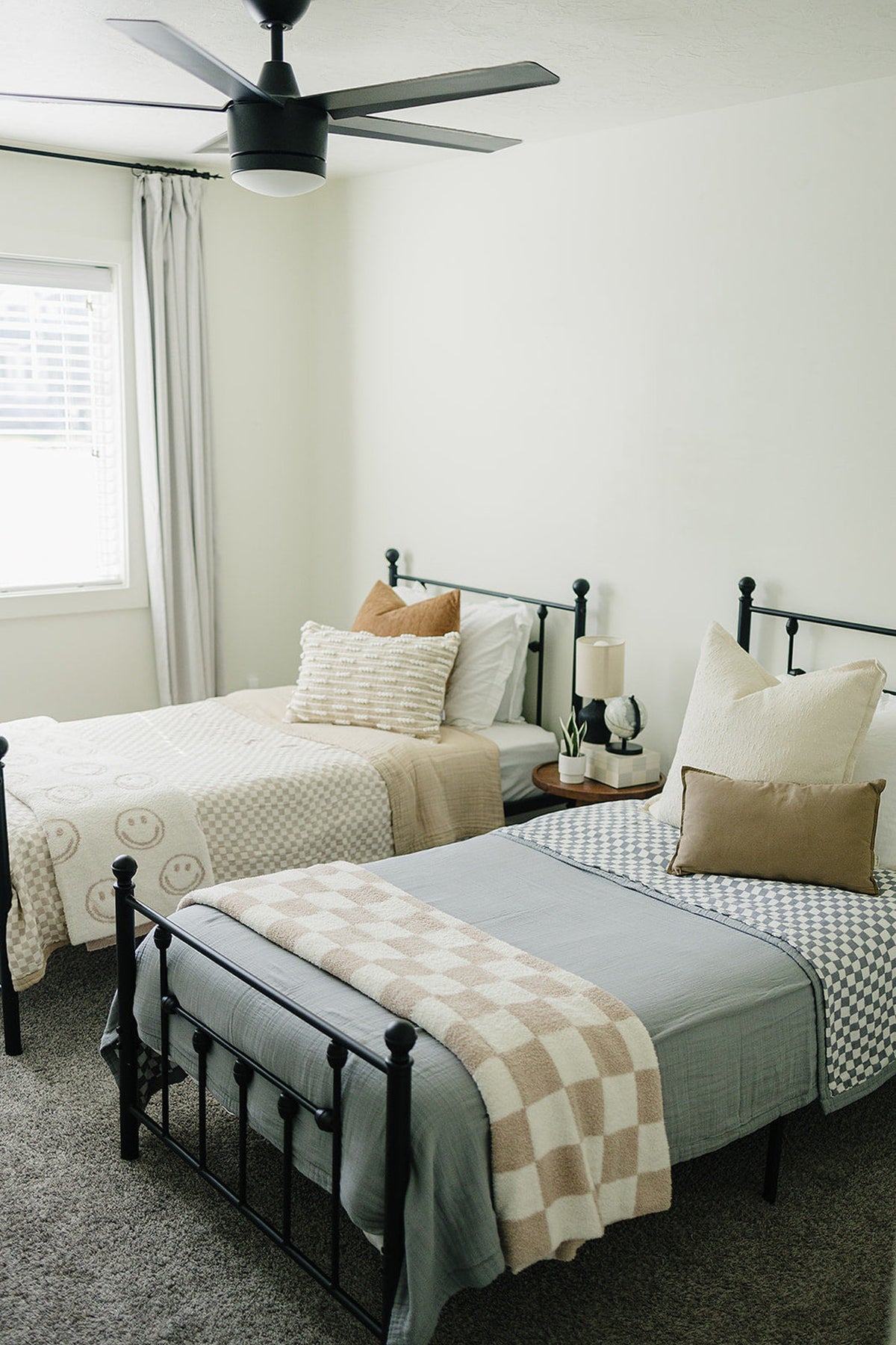 Two twin beds with black metal frames feature taupe checkered throws and decorative pillows in a bright bedroom.