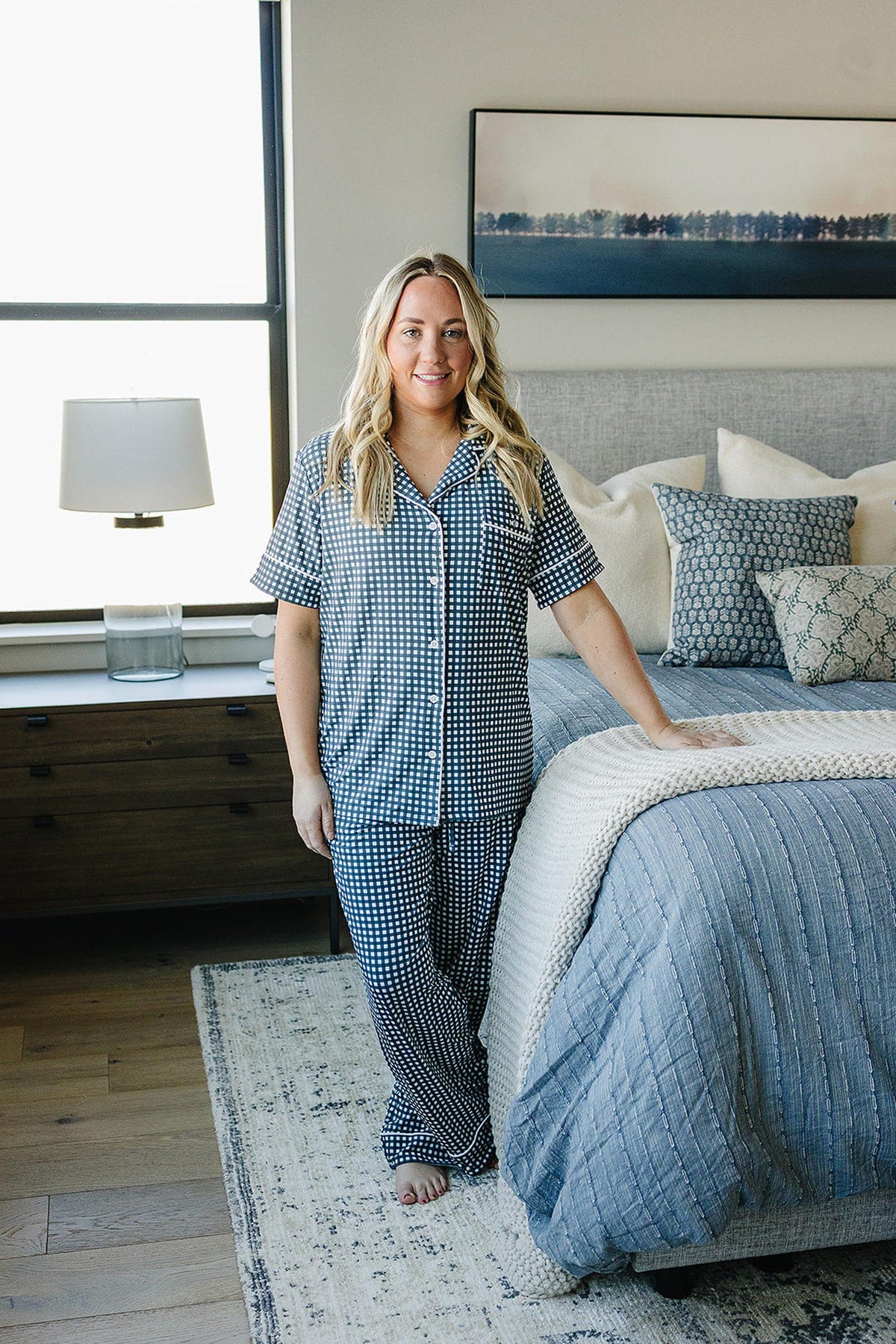 A woman in navy gingham pajamas stands beside a bed with patterned pillows in a bright bedroom.