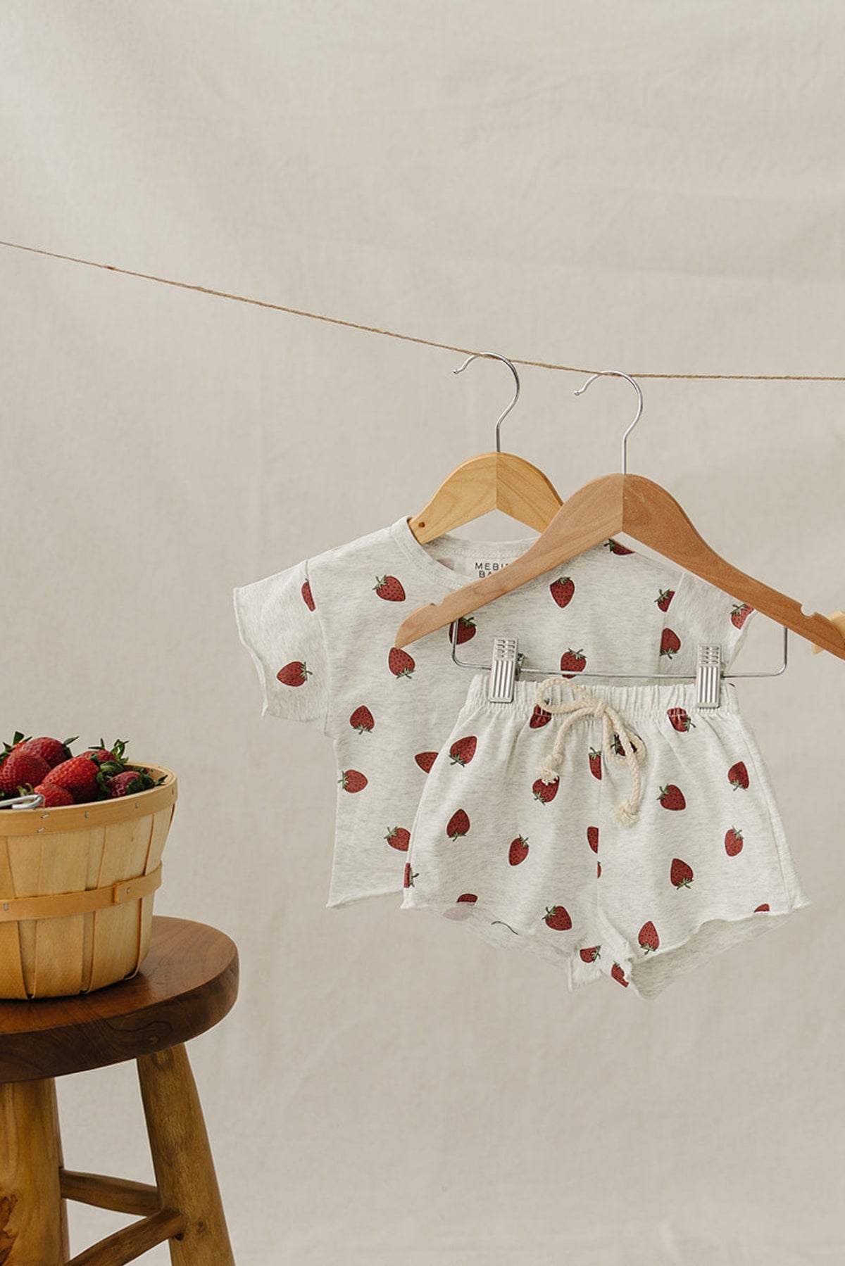 Light grey cotton shorts and t-shirt set with strawberry prints, displayed on wooden hangers next to a basket of strawberries.