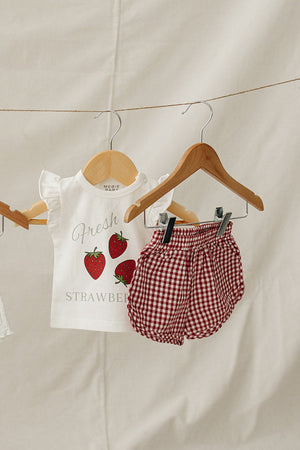 A white cotton t-shirt featuring a playful strawberry graphic hangs on a wooden hanger alongside red gingham shorts, set against a neutral fabric backdrop. Ideal for springtime wear.