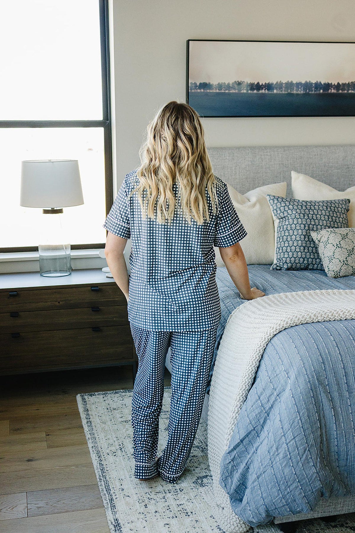 A woman in a navy gingham pajama set stands by a bed with blue and white bedding.