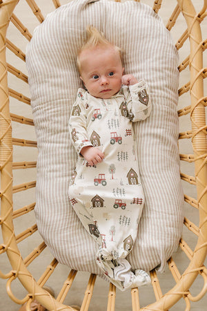 A baby lies on a striped cushion in a woven bassinet, wearing a soft, cream-colored bamboo gown adorned with playful farm-themed prints, perfect for ages 0-3 months.