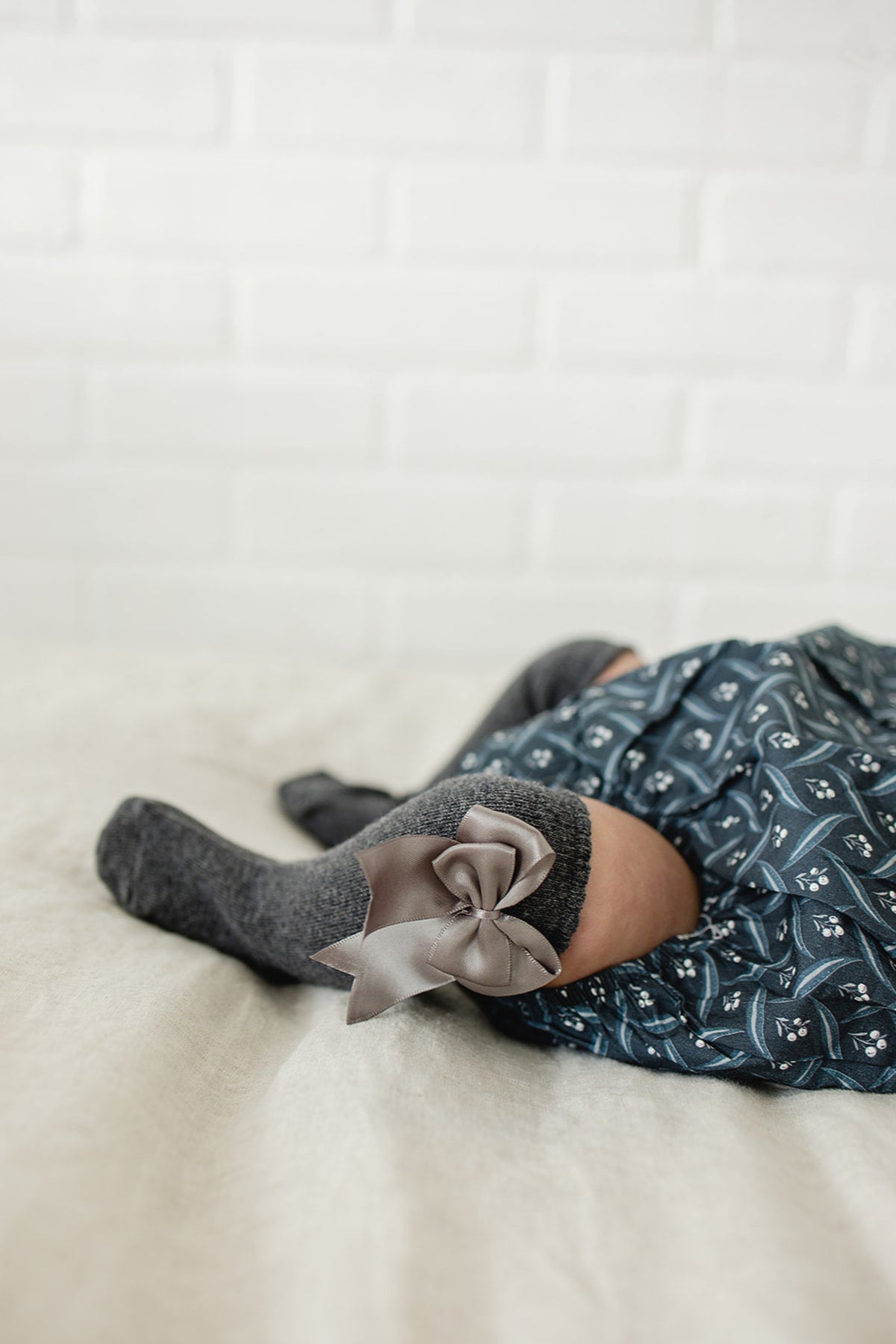A baby wearing gray high-rise socks with a satin bow, paired with a patterned blue dress, on a beige surface.