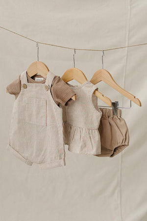 Three baby outfits are displayed on wooden hangers against a soft beige background, featuring earthy tones and subtle stripes.