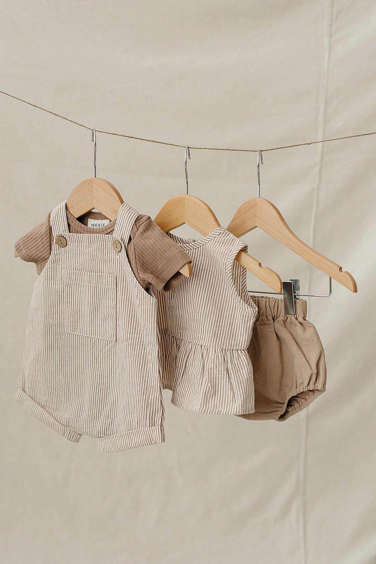 Three pieces of baby clothing hang on wooden hangers against a neutral backdrop, featuring tan stripes and a peplum hem.