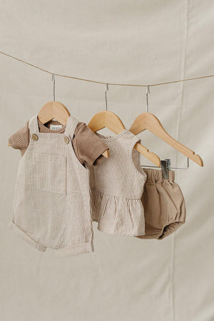 Three pieces of baby clothing hang on wooden hangers against a neutral backdrop, featuring tan stripes and a peplum hem.
