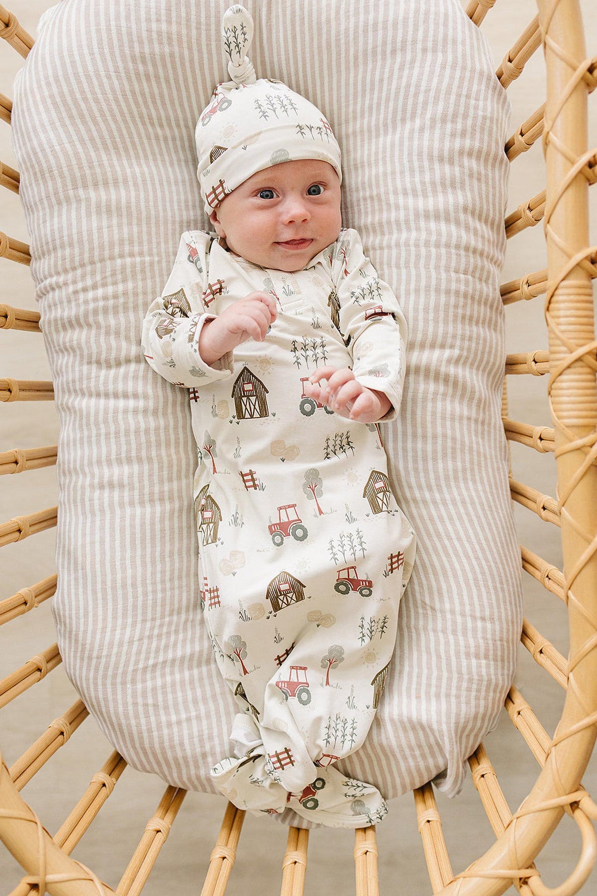 A baby in a soft, cream-colored bamboo gown with a playful farm design smiles while lying in a woven bassinet. The gown features fold-over hands and a matching hat.