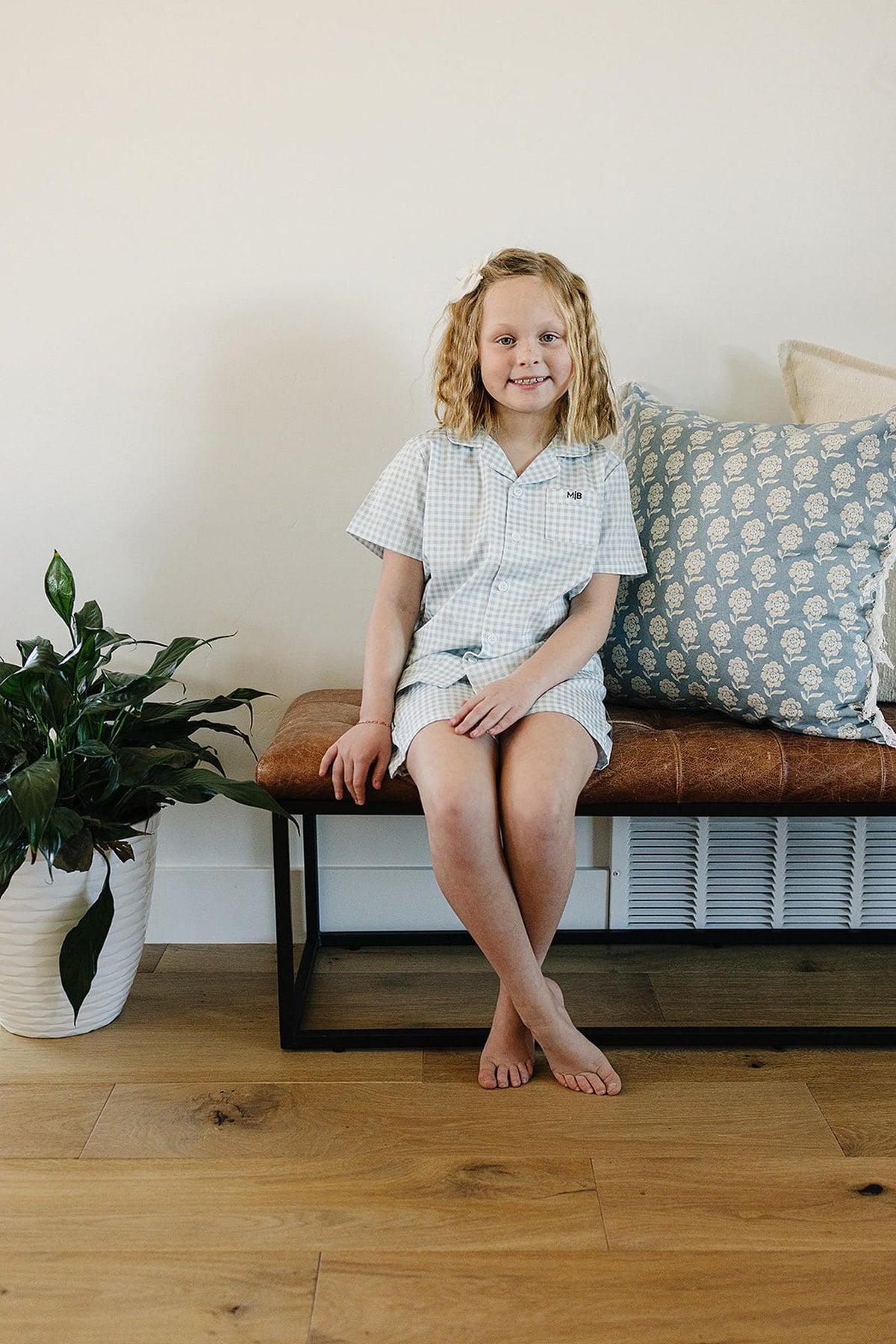A child wearing a light blue gingham pajama set sits on a bench beside a potted plant and cushions.