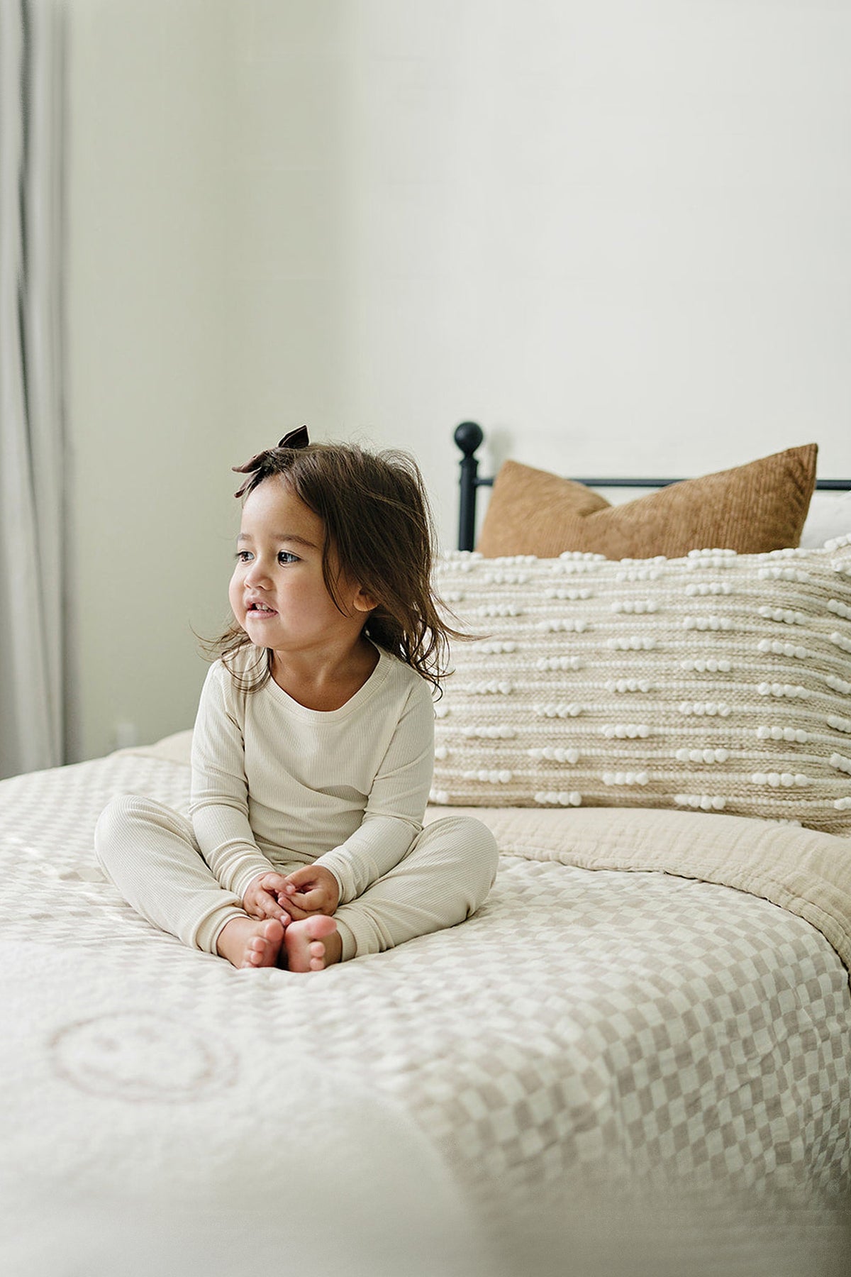 A child in cream pajamas sits on a bed with a taupe checkered quilt and textured pillows.