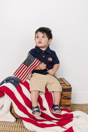A young child sits on a wooden crate, draped in a red, white, and blue blanket. He wears a navy polo shirt with a flag detail, paired with beige shorts.