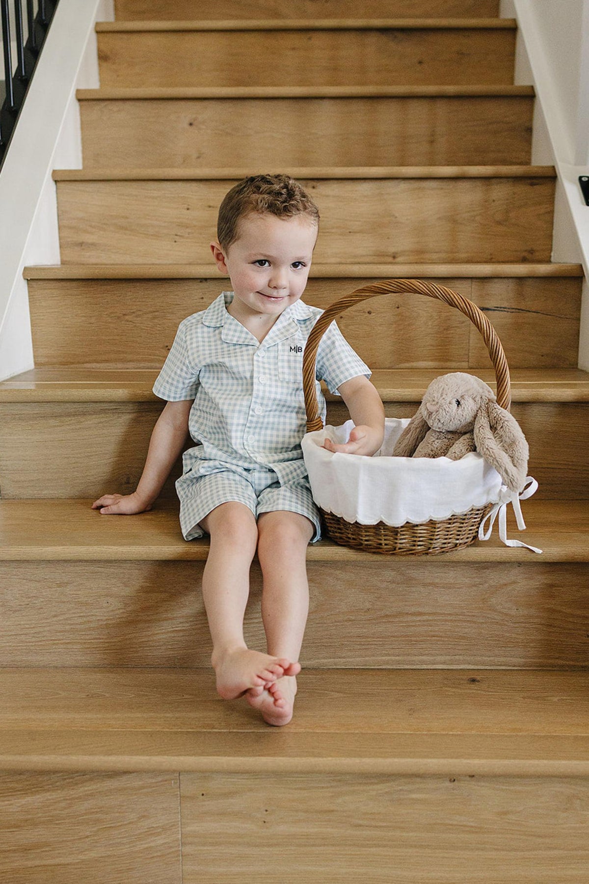 A child in light blue gingham pajamas sits on wooden stairs beside a basket with a stuffed rabbit.