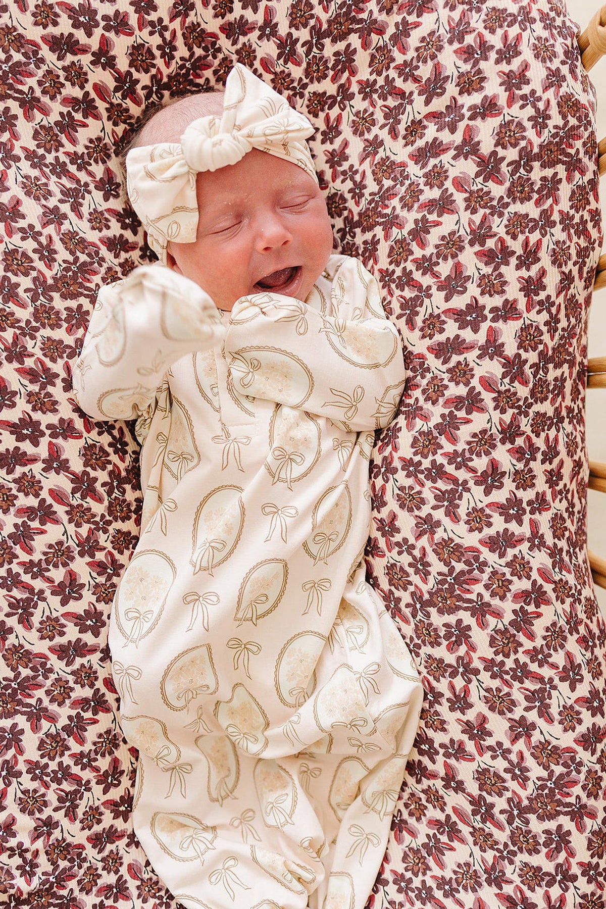 A baby in a vintage bouquet bamboo knot gown, featuring a soft, cream fabric with a floral pattern, is peacefully resting on a floral-patterned blanket with a matching headband.