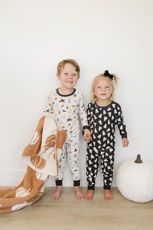 Two young children in Halloween-themed pajamas, one holding a pumpkin-patterned blanket, stand next to a white pumpkin.