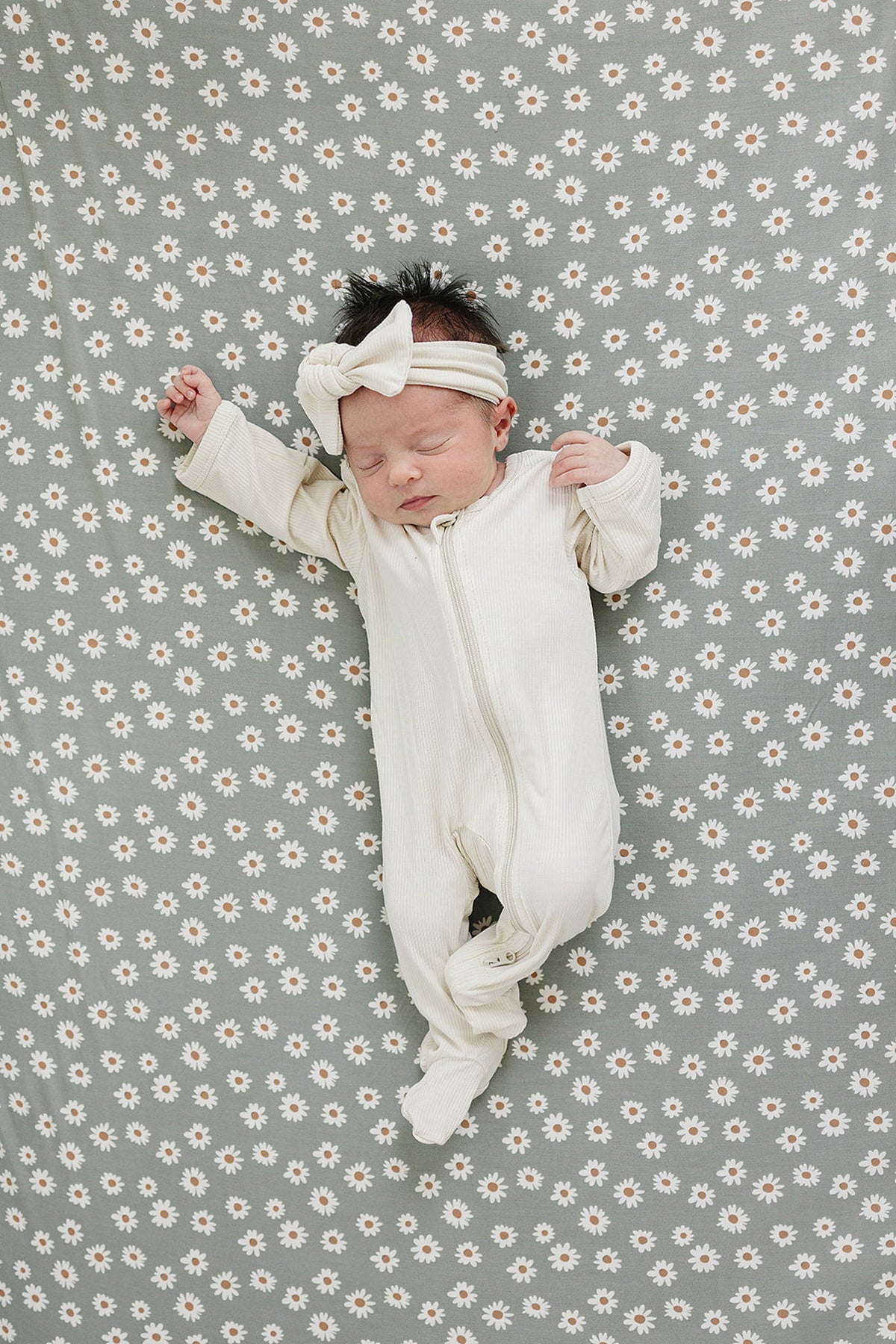 A peaceful newborn wearing a cream-colored onesie and headband lies on a light green daisy-patterned crib sheet.
