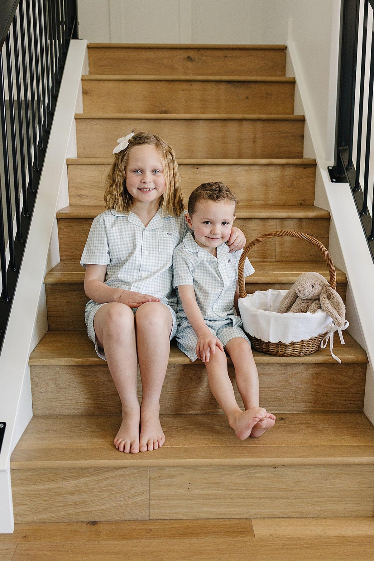 Two children in light blue gingham pajamas sit on wooden stairs with a wicker basket holding a plush toy.