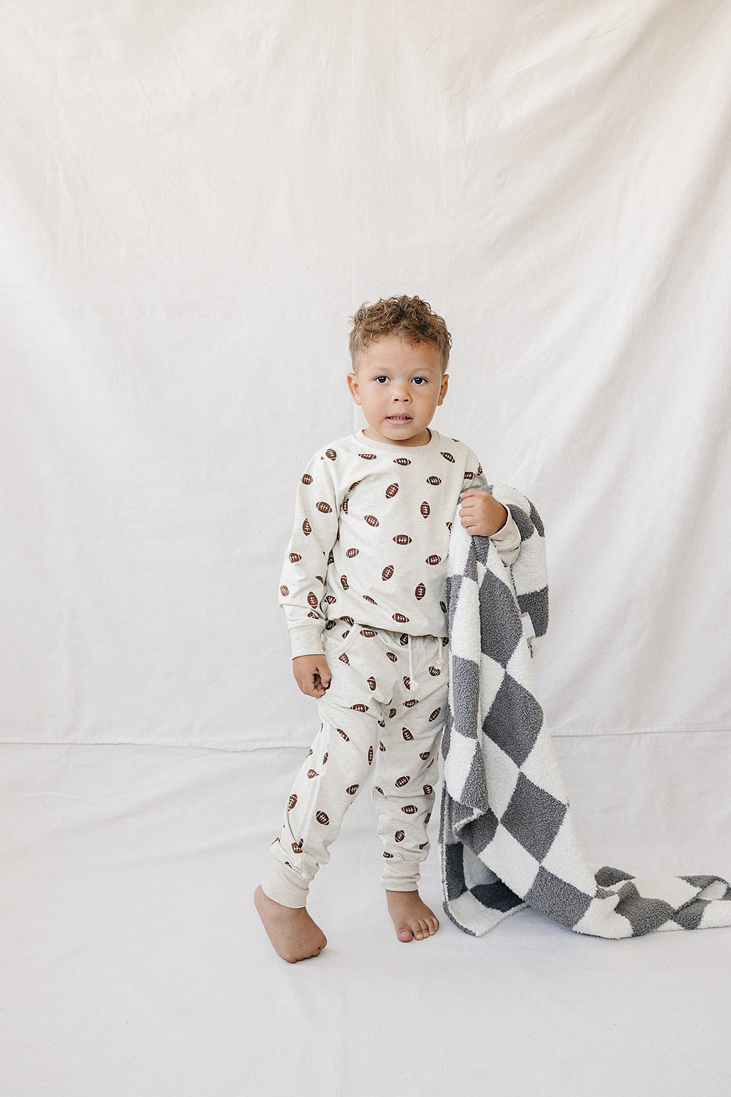 A young child stands barefoot, wearing a cream-colored football print French terry cotton set. They hold a cozy gray and white checkered blanket against a neutral backdrop.