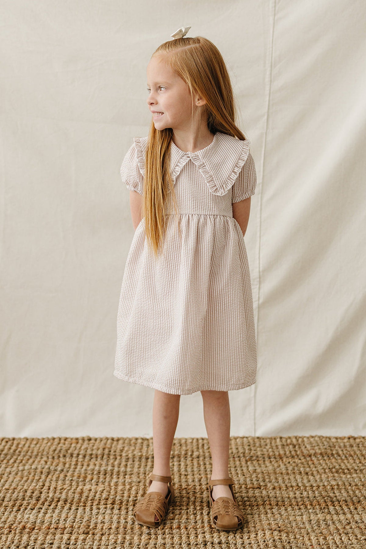 A girl with long red hair smiles while wearing a taupe striped cotton dress with a Peter Pan collar.