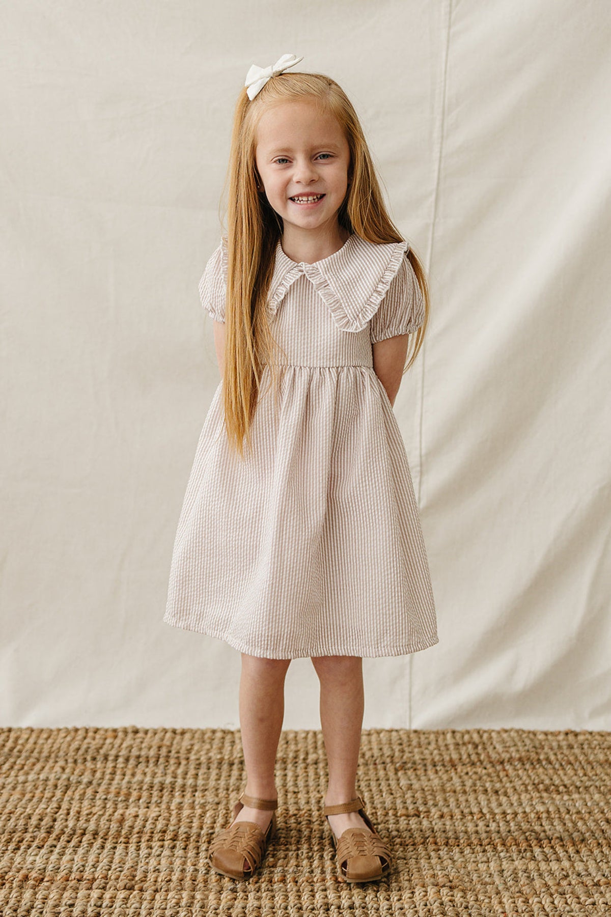 A smiling girl wears a taupe striped cotton dress with a Peter Pan collar, standing on a textured woven rug.